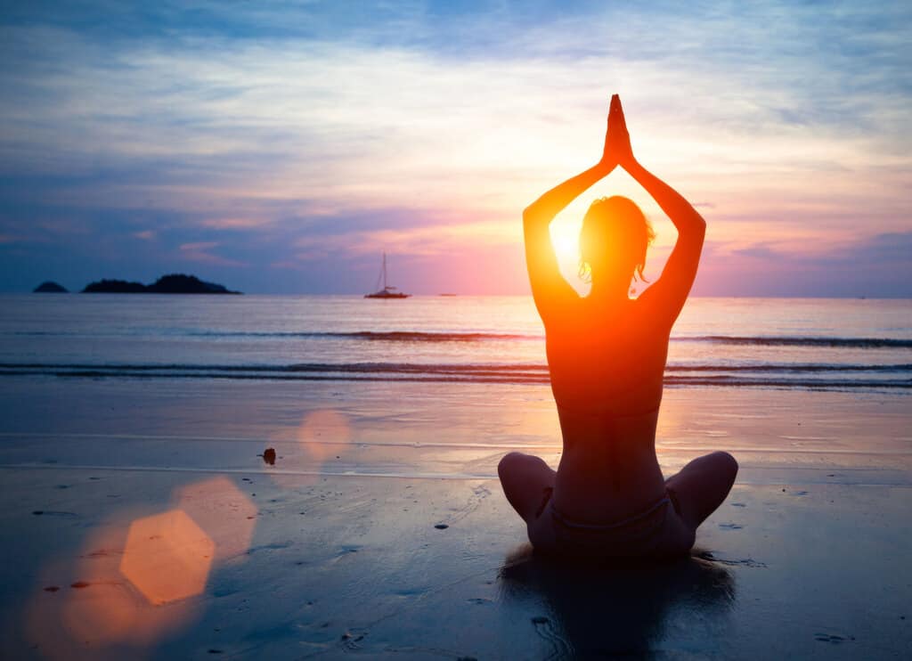 woman at sunset on beach in easy cross leg asana with warrior 1 hands