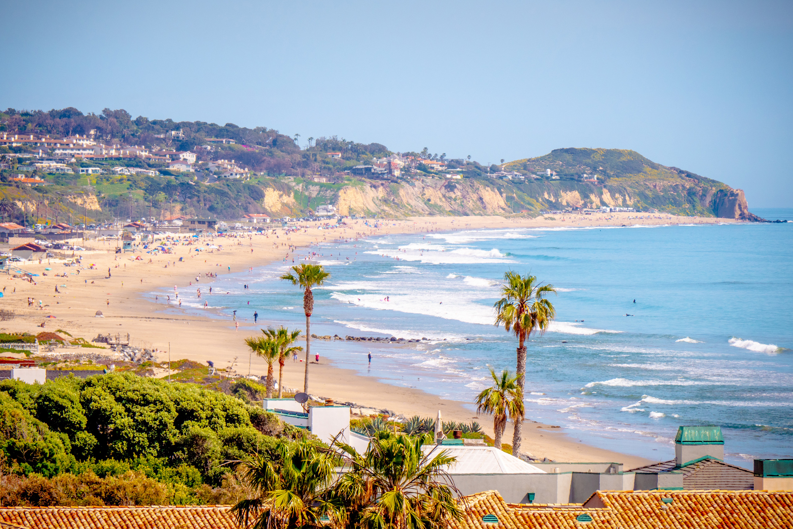 Crowded Malibu beach with waves and coastal homes.