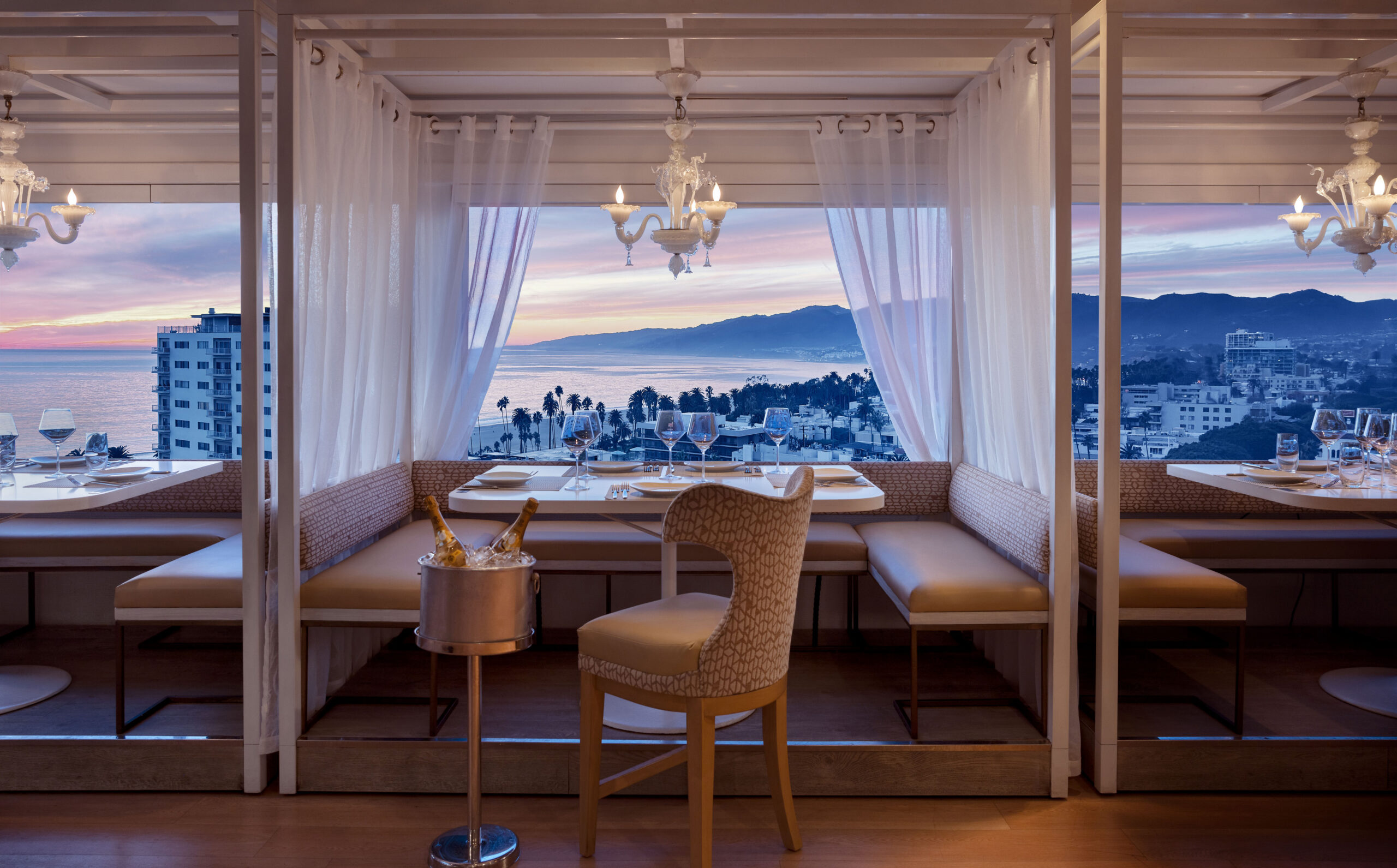Elegant restaurant booth with ocean sunset view, white curtains, and table set for dining