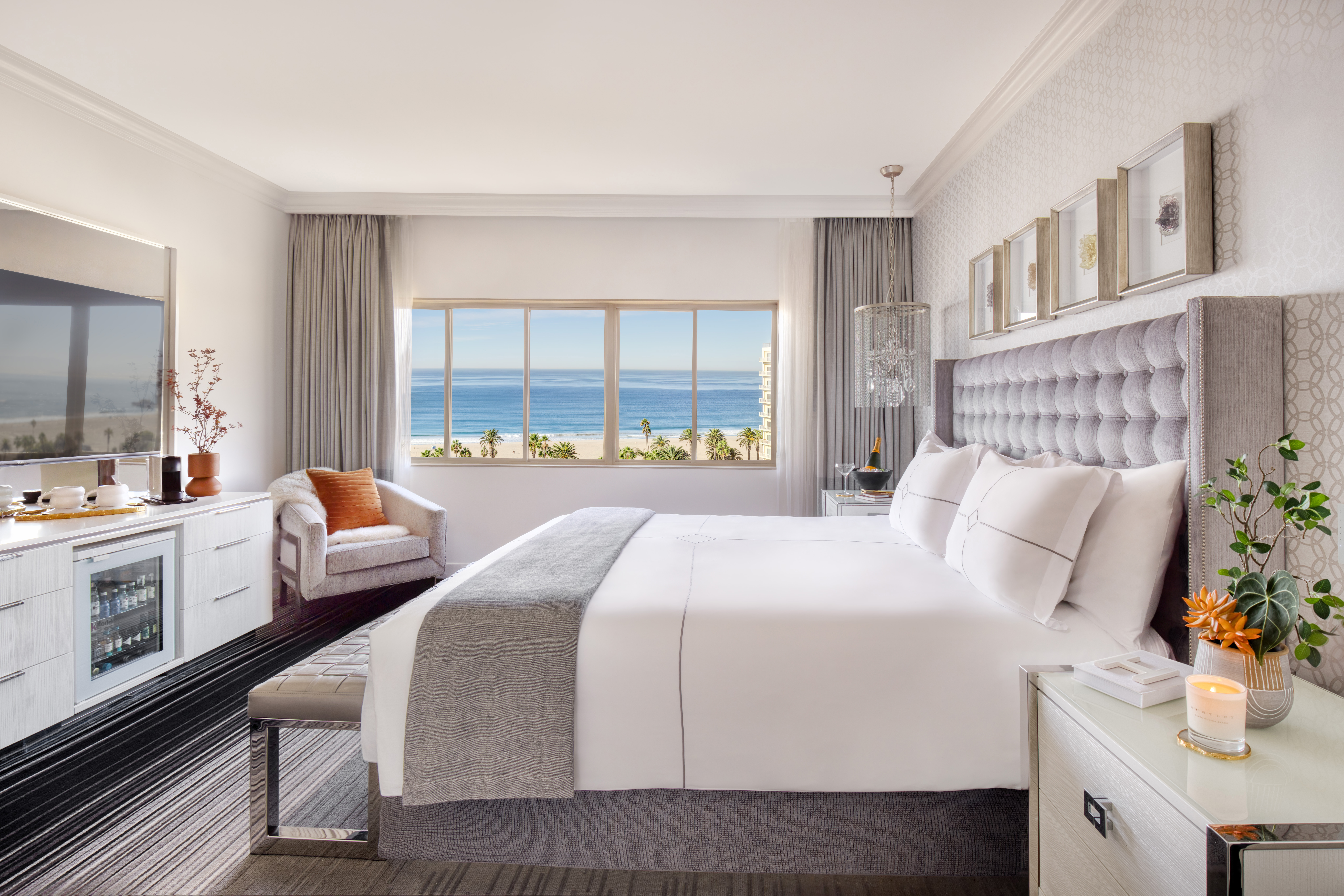 Bright coastal hotel room with large bed, TV console, and ocean view through wide window
