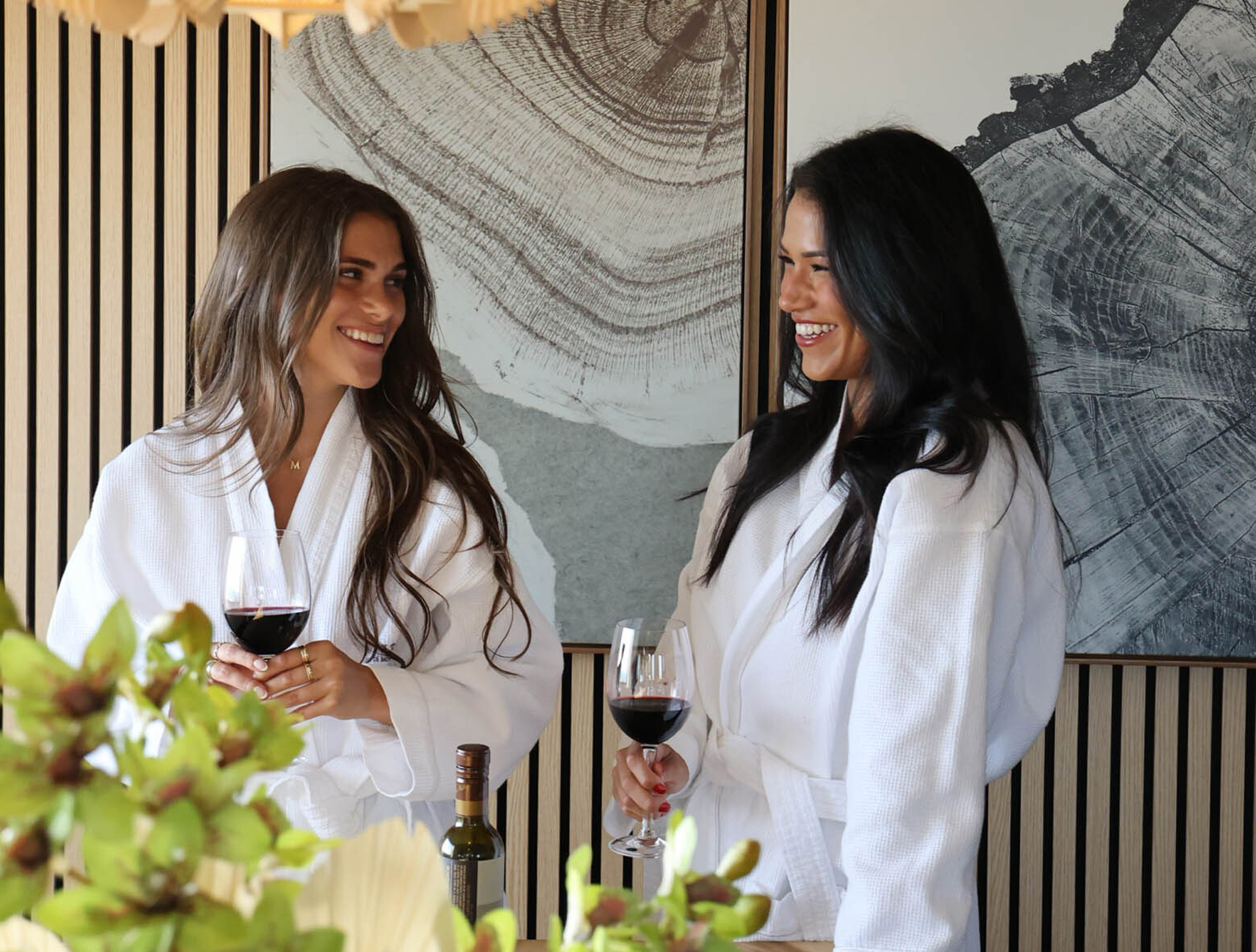 Two women in white robes smiling and holding glasses of red wine in a modern interior