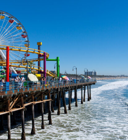 Santa Monica Pier extending over the ocean, featuring a colorful Ferris wheel and roller coaster beside a sunny California beach.