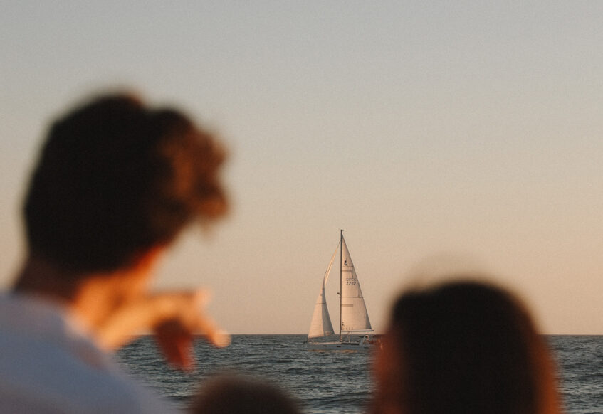 Blurred family in the foreground looking toward a sailboat on the ocean at sunset.