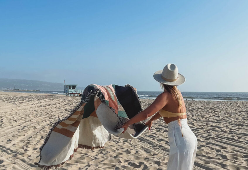 Person shaking out a patterned blanket on a sandy beach near the shoreline.