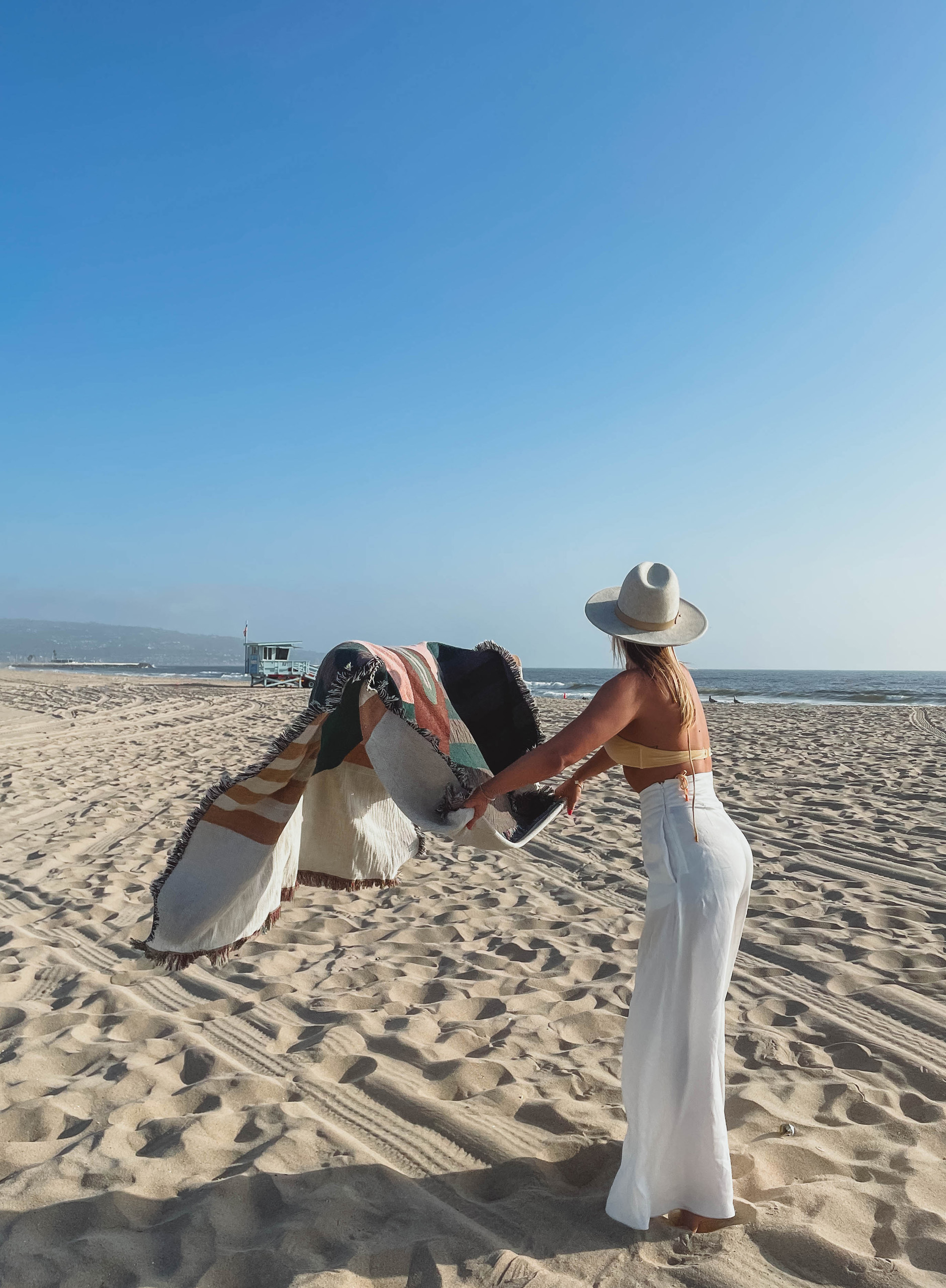 Person shaking out a patterned blanket on a sandy beach near the shoreline.