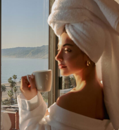 Woman in a white robe with a towel wrapped around her hair holds a mug by a hotel window overlooking the ocean and palm-lined beach.