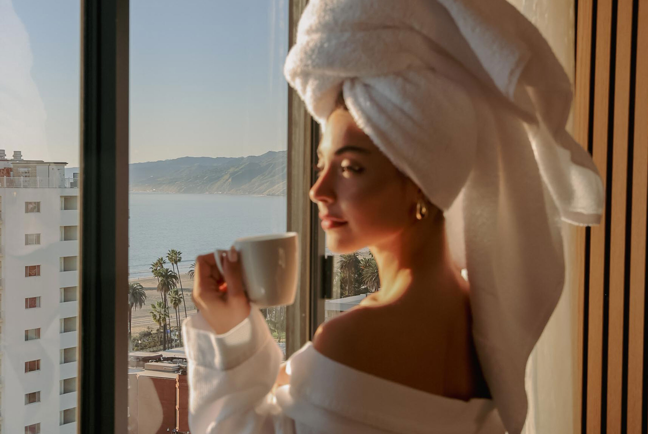 Woman in a white robe with a towel wrapped around her hair holds a mug by a hotel window overlooking the ocean and palm-lined beach.