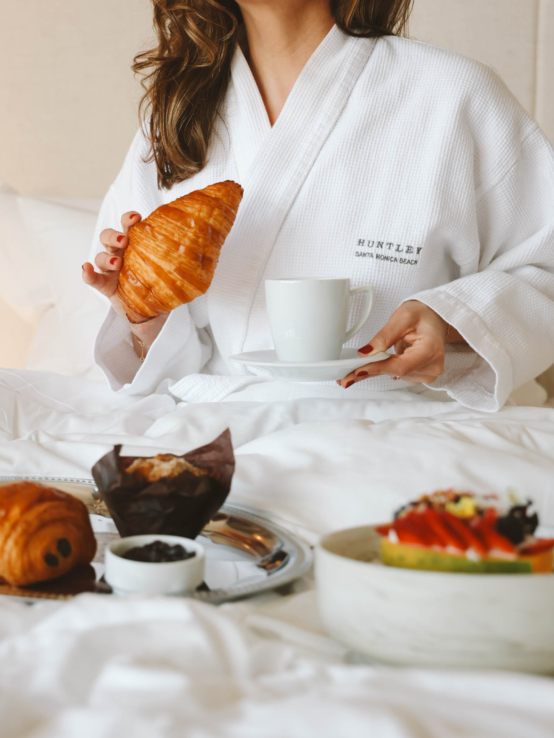 Alt text: Woman in a white Huntley Santa Monica Beach robe enjoying a croissant and coffee with breakfast served in bed.