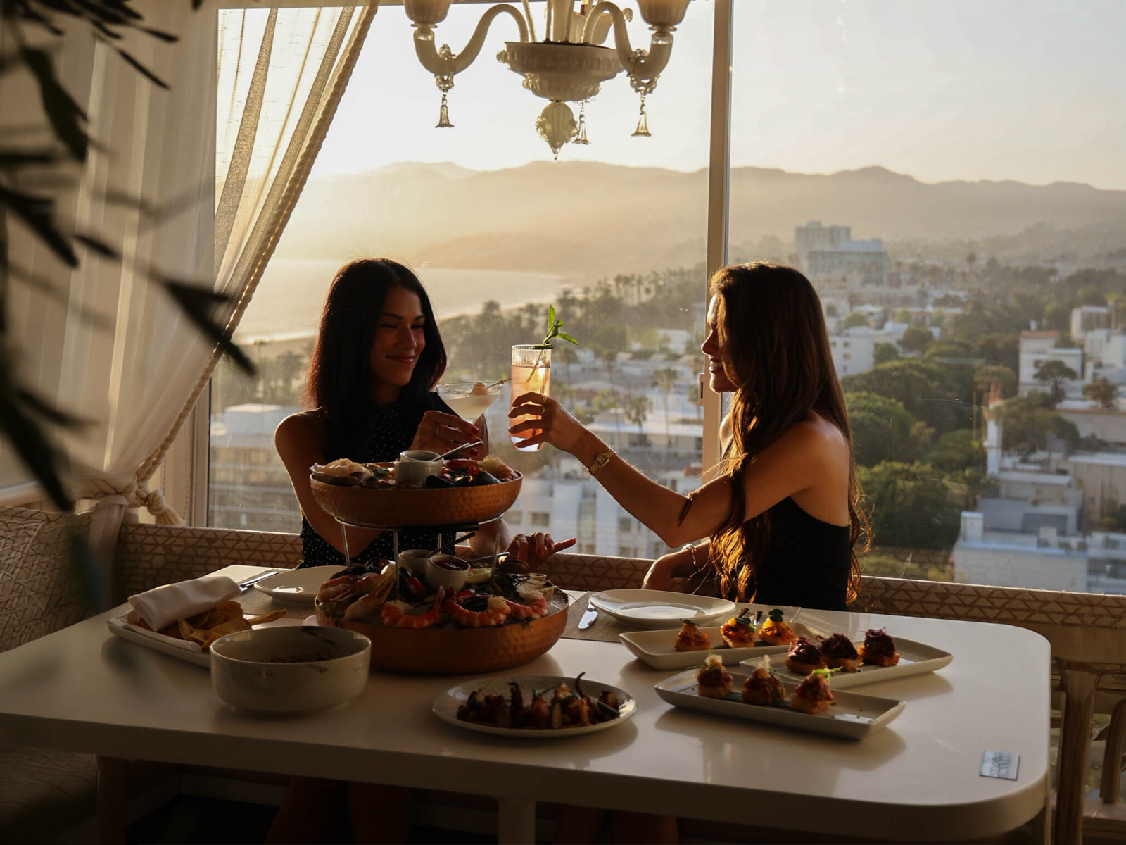 Two women toast drinks over a seafood meal with ocean and city views at sunset.