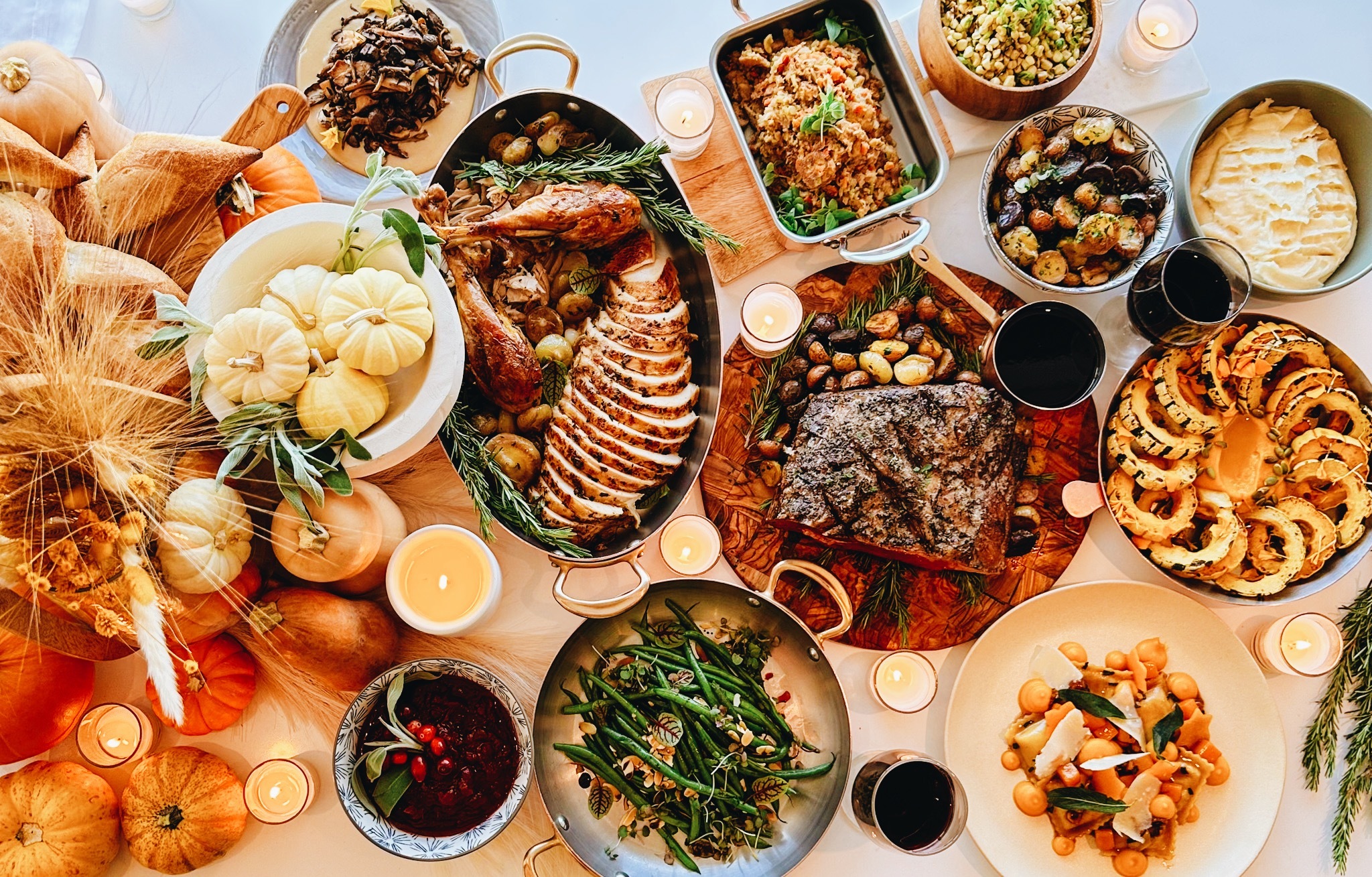 Overhead view of a Thanksgiving feast with roasted turkey, meat, vegetables, side dishes, pumpkins, and candles.