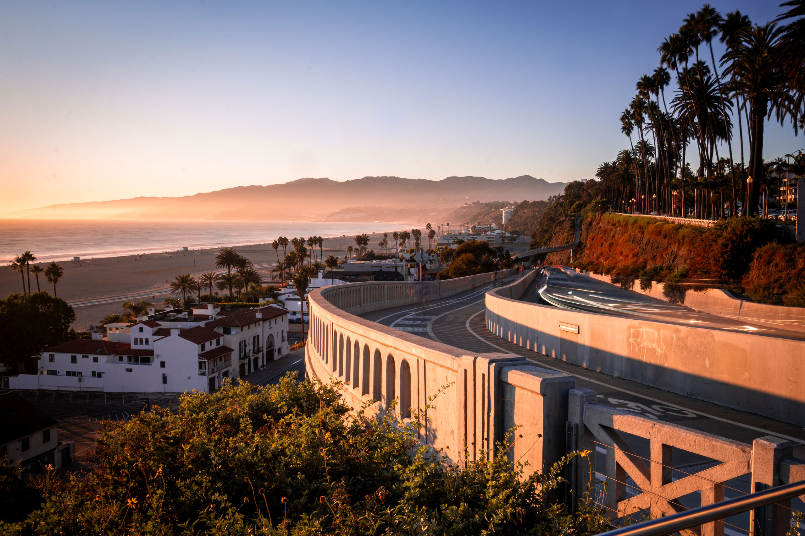 Coastal highway curving above a palm-lined beach in Santa Monica at sunset, with mountains in the distance.
