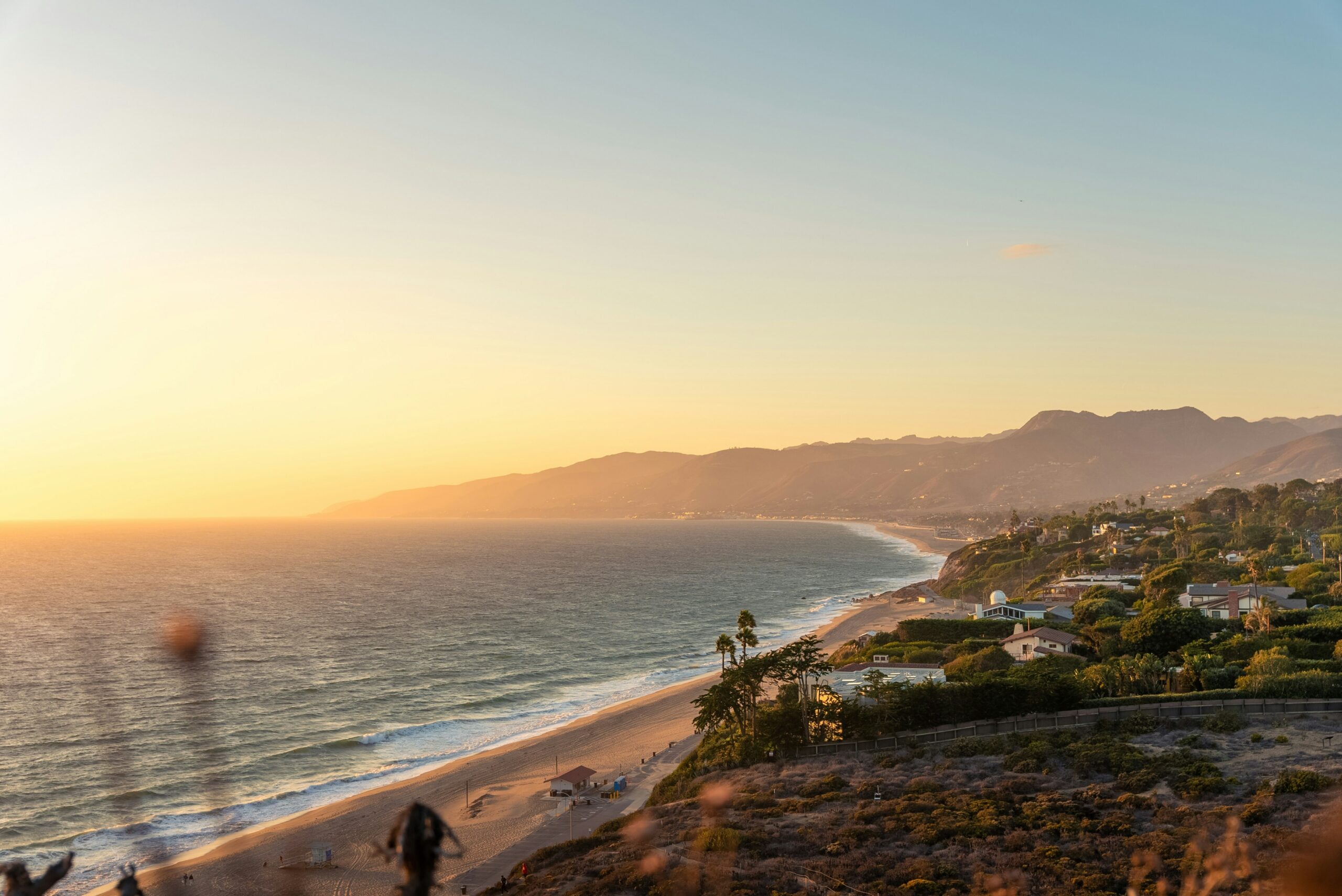 Coastal beach and hillside homes overlooking the ocean at sunset, with mountains in the distance.