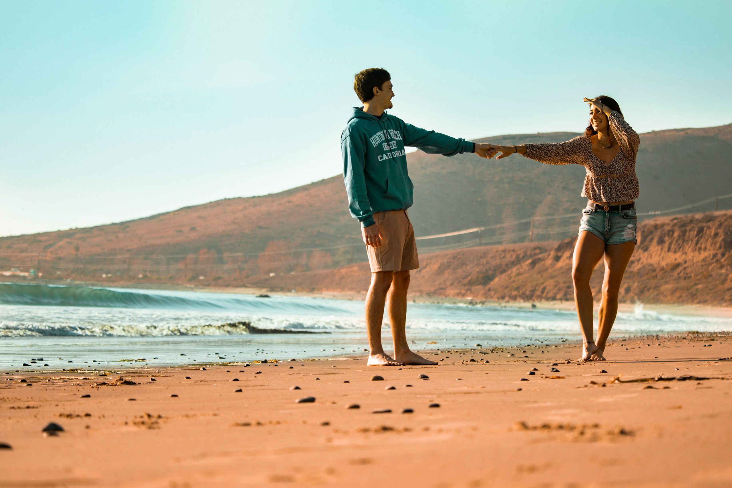 Couple holding hands while walking barefoot on a sandy beach with coastal hills in the background.